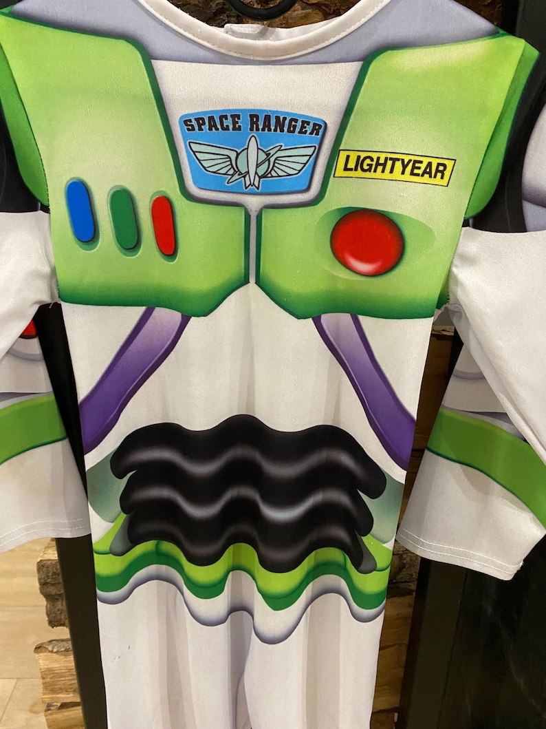 May include: A Buzz Lightyear costume featuring a white and green design with the words "SPACE RANGER" and "LIGHTYEAR". The costume has a green collar and accents, with a red button and three colored buttons on the chest.