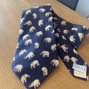 Op de afbeelding: Marineblauwe stropdas met een herhalend patroon van beige olifanten. De stropdas is gemaakt van een gladde stof en ligt op een houten oppervlak. Het label luidt "MADE IN THAILAND DRY CLEAN ONLY".