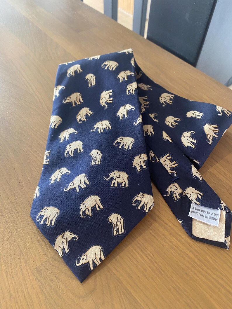 Op de afbeelding: Marineblauwe stropdas met een herhalend patroon van beige olifanten. De stropdas is gemaakt van een gladde stof en ligt op een houten oppervlak. Het label luidt "MADE IN THAILAND DRY CLEAN ONLY".