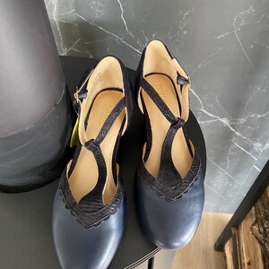 May include: A pair of navy blue T-strap shoes with a rounded toe and a scalloped edge detail along the vamp. The shoes feature a textured black strap design, a buckle closure at the ankle, and a tan interior lining with the Hotter brand name visible.