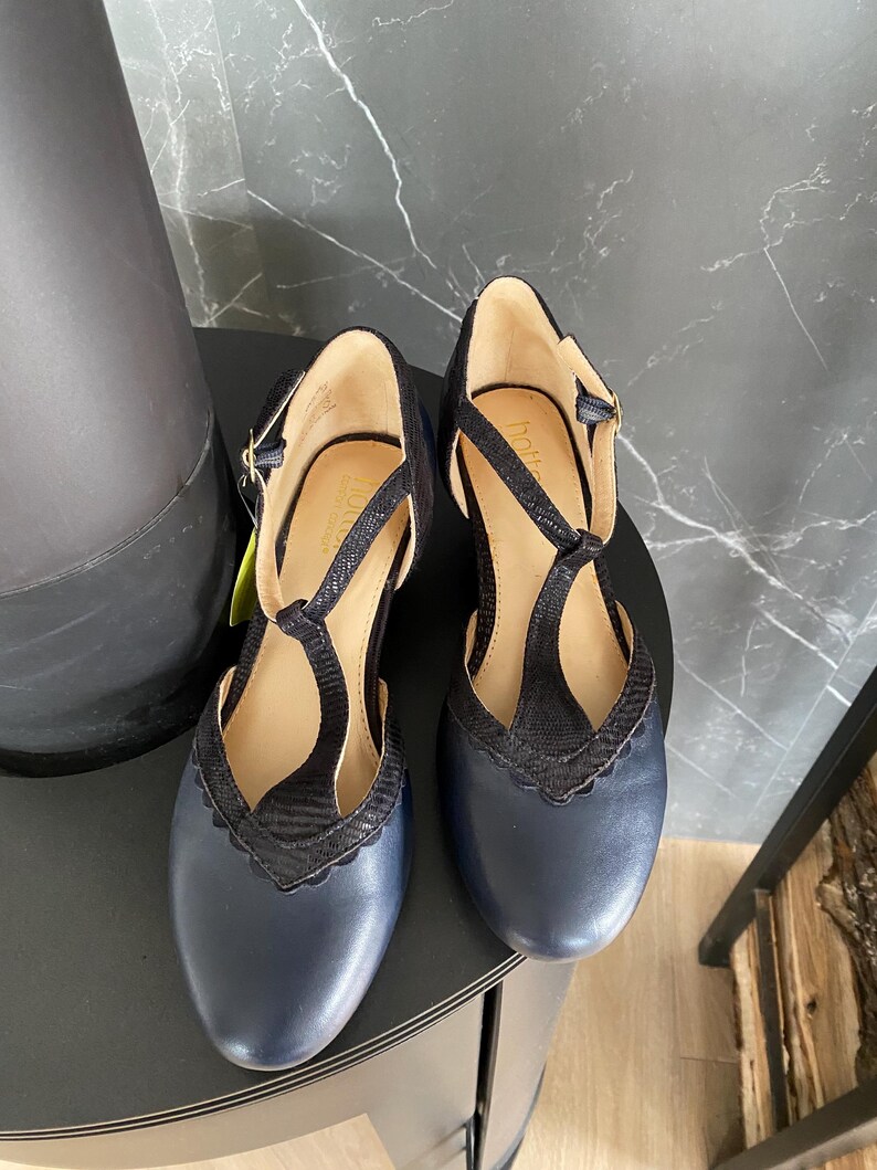 May include: A pair of navy blue T-strap shoes with a rounded toe and a scalloped edge detail along the vamp. The shoes feature a textured black strap design, a buckle closure at the ankle, and a tan interior lining with the Hotter brand name visible.