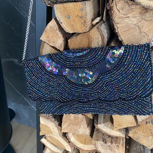 May include: A black beaded clutch with a silver chain strap. The clutch is decorated with iridescent sequins and beads in shades of blue, purple, and green. The bag is rectangular and appears to be made of a soft fabric.
