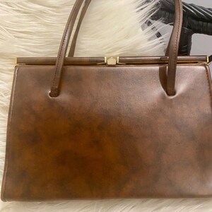 Könnte beinhalten: Eine braune Vintage-Handtasche mit rechteckiger Form und zwei Henkeln. Die Tasche hat eine strukturierte Oberfläche und goldfarbene Akzente am Verschluss. Die Henkel sind dünn und aus dem gleichen Material wie die Tasche. Die Tasche liegt auf einer weißen, flauschigen Oberfläche.