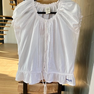 Puede incluir: Una blusa blanca de manga corta con cuello cuadrado y estampado de rayas verticales. La blusa tiene un cordón en la cintura y un dobladillo con volantes. La marca "Woodland Active" es visible en texto azul en el dobladillo.