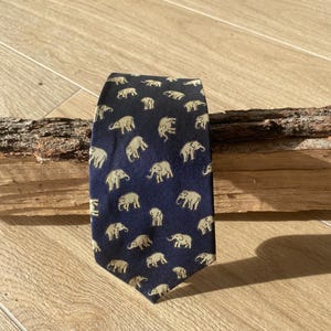 Op de afbeelding: Marineblauwe stropdas met een herhaald patroon van beige olifanten. De stropdas is gemaakt van een gladde stof en wordt gepresenteerd op een houten oppervlak. De olifanten worden in verschillende poses afgebeeld.