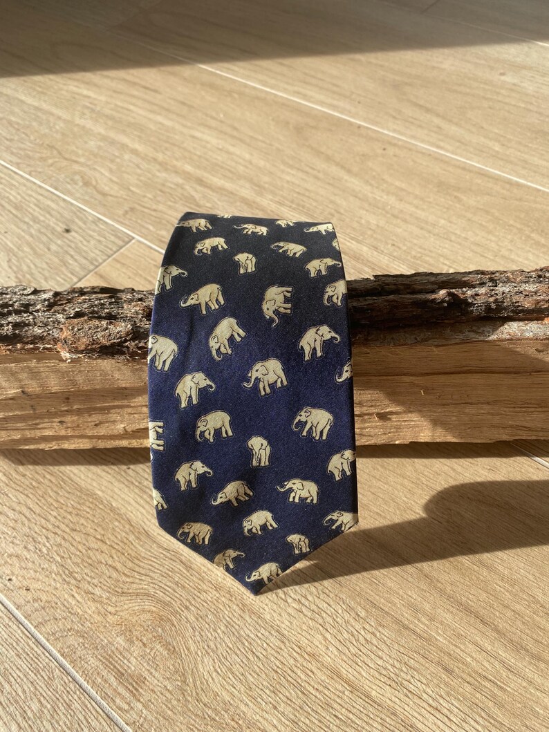 Op de afbeelding: Marineblauwe stropdas met een herhaald patroon van beige olifanten. De stropdas is gemaakt van een gladde stof en wordt gepresenteerd op een houten oppervlak. De olifanten worden in verschillende poses afgebeeld.