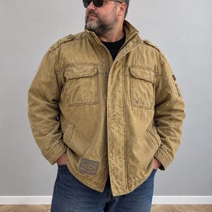 May include: A tan waxed cotton field jacket featuring a high collar, shoulder epaulets, and two chest pockets with flaps. The jacket includes a front zipper closure, a small logo on the left chest, and a rectangular patch on the lower right front.