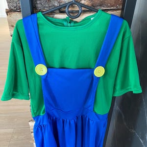 May include: A green short-sleeved shirt with blue overalls, featuring large yellow buttons. The costume is hanging on a black hanger against a wood and black background. The outfit is designed for a child.