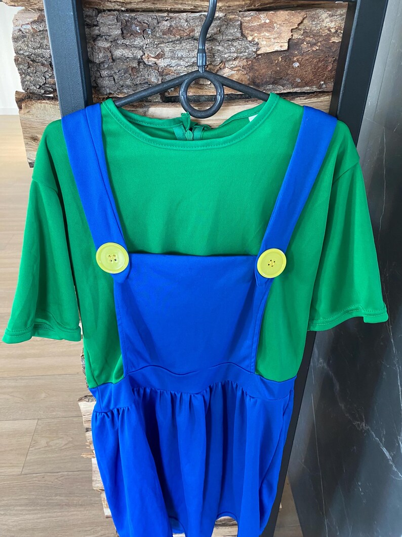 May include: A green short-sleeved shirt with blue overalls, featuring large yellow buttons. The costume is hanging on a black hanger against a wood and black background. The outfit is designed for a child.