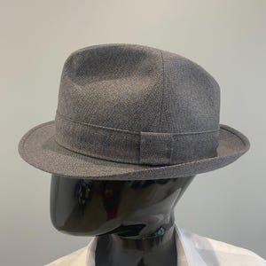 Pode incluir: Um chapéu fedora cinzento com uma coroa estruturada e uma aba larga. O chapéu apresenta uma faixa de gorgorão ao redor da base da coroa. O chapéu é exibido em uma cabeça de manequim preta, com uma camisa branca visível.