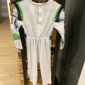 May include: White Buzz Lightyear costume with green, black, and purple accents. The costume features long sleeves and a jumpsuit design, with details mimicking the character's space suit. The costume is hanging on a black hanger.