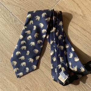 Op de afbeelding: Marineblauwe stropdas met een herhaald patroon van beige olifanten. De stropdas is gemaakt van een gladde stof en ligt op een houten oppervlak. De olifanten staan in verschillende poses.