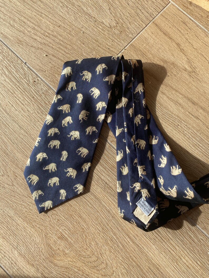 Op de afbeelding: Marineblauwe stropdas met een herhaald patroon van beige olifanten. De stropdas is gemaakt van een gladde stof en ligt op een houten oppervlak. De olifanten staan in verschillende poses.