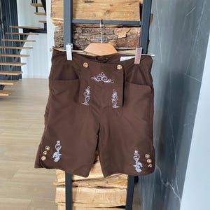 May include: Brown lederhosen shorts with decorative white embroidery and gold-colored buttons. The shorts have two front pockets and are hanging on a wooden hanger. A traditional garment, often worn during festivals.