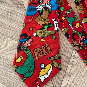 Könnte beinhalten: Rote Krawatte mit Goofy-Illustrationen in sportlichen Posen. Die Krawatte trägt die Aufschrift "FORE" und "10 LBS" sowie Golf- und Gewichtheber-Motive. Die Krawatte ist mit Sternen und anderen festlichen Elementen verziert.
