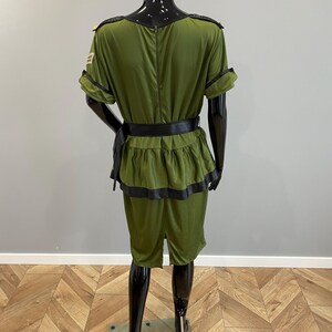 May include: An olive green costume with black trim and a matching belt. The outfit features a short-sleeved top with epaulettes and shorts. The costume is displayed on a black mannequin.