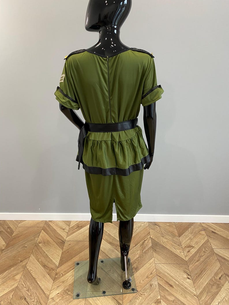 May include: An olive green costume with black trim and a matching belt. The outfit features a short-sleeved top with epaulettes and shorts. The costume is displayed on a black mannequin.