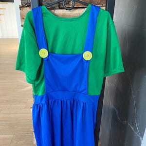 May include: A green t-shirt with blue overalls attached, featuring large yellow buttons. The costume is hanging on a black hanger. The overall dress has a gathered skirt.