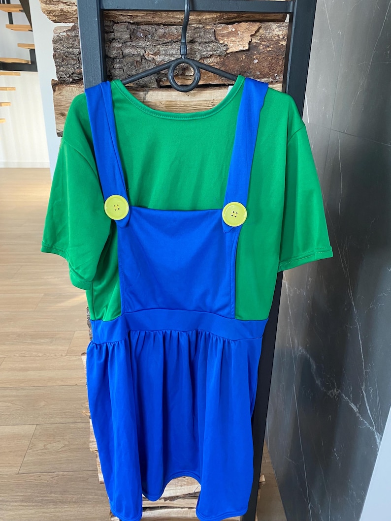 May include: A green t-shirt with blue overalls attached, featuring large yellow buttons. The costume is hanging on a black hanger. The overall dress has a gathered skirt.