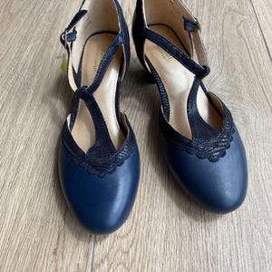 May include: A pair of navy blue T-strap heels featuring a smooth leather-like toe box and a textured, snakeskin-patterned black overlay with a scalloped edge. The shoes include an adjustable ankle strap with a metal buckle and a low block heel.