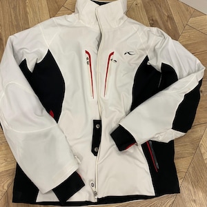 Może przedstawiać: Biała, czarna i czerwona kurtka narciarska z wysokim kołnierzem i kieszeniami zapinanymi na zamek. Kurtka ma czarne panele po bokach i na rękawach, z czerwonymi akcentami na zamkach i mankietach. Logo jest widoczne na piersi.