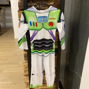 May include: A Buzz Lightyear costume, featuring a white jumpsuit with green and purple accents. The chest area has a green panel with the words "SPACE RANGER" and "LIGHTYEAR". The costume has long sleeves and is hanging on a black metal rack.