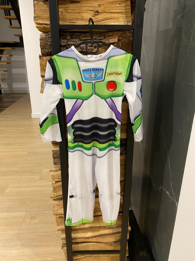 May include: A Buzz Lightyear costume, featuring a white jumpsuit with green and purple accents. The chest area has a green panel with the words "SPACE RANGER" and "LIGHTYEAR". The costume has long sleeves and is hanging on a black metal rack.