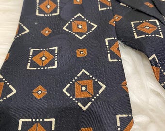 Puede incluir: Corbata azul marino con un patrón geométrico repetido de diamantes marrones y cuadrados punteados blancos. La corbata está hecha de una tela texturizada y es adecuada para vestimenta formal o de negocios.