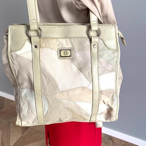 May include: Cream leather tote bag with a patchwork design. The bag has a rectangular shape, two top handles, and a small rectangular emblem. The patchwork is made up of various shades of cream and beige, creating a unique pattern.