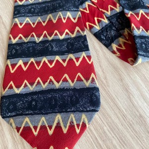 Peut inclure: Une cravate présentant un motif répétitif de bandes horizontales rouges, gris foncé et gris clair. Le design comprend de fines lignes en zigzag jaune doré superposées sur les sections rouges et des bandes gris foncé avec un motif circulaire texturé.