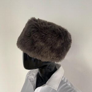 May include: A dark brown faux fur hat with a rectangular shape. The hat is displayed on a black mannequin head, paired with a white collared shirt. The fur appears soft and plush.