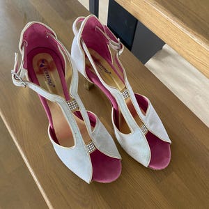 May include: A pair of silver and pink open-toe high heels with a strap and buckle closure. The heels have a glittery finish and are embellished with small rhinestones. The interior is a vibrant pink, and the shoes are displayed on a wooden surface.