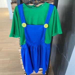 May include: A green and blue costume dress with yellow button accents. The dress features a green short-sleeved top and blue overalls with a pleated skirt. The costume is hanging on a black hanger.