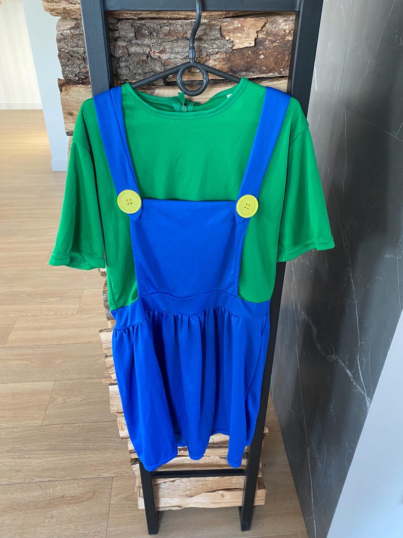 May include: A green and blue costume dress with yellow button accents. The dress features a green short-sleeved top and blue overalls with a pleated skirt. The costume is hanging on a black hanger.
