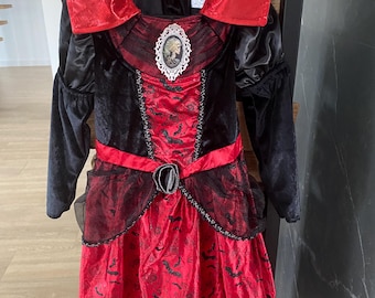 May include: A red and black vampire costume with a high collar and long sleeves. The dress features a red skirt with a bat pattern and a black bodice with a decorative brooch. The costume is hanging on a black hanger.