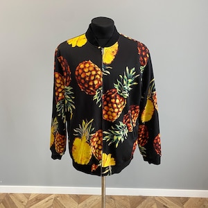May include: Black bomber jacket with a repeating pineapple design. The jacket has a black collar and cuffs, and a zip. The pineapple pattern features yellow, green, and brown colours, creating a tropical theme.