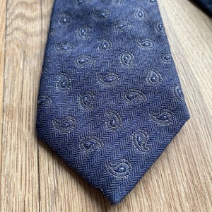 May include: A navy blue tie with a paisley pattern. The tie has a textured, woven appearance with a herringbone weave. The paisley design is a lighter shade of blue, creating a subtle contrast. The tie is a classic accessory.