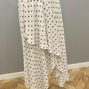 May include: A white midi skirt with a navy blue waistband and a repeating pattern of small, dark blue floral shapes. The skirt has a layered, asymmetrical design and is displayed on a metal stand.