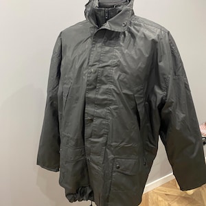 以下が含まれることがあります： ハイカラーと複数のポケットが付いた、濃いグレーの防水ジャケット。 ジャケットはジッパー開閉とボタンのディテールを備えています。裾にはドローストリングが付いています。 ジャケットはマネキンに展示されています。