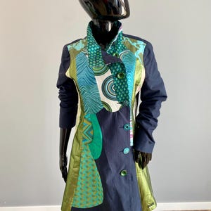 Pode incluir: Um casaco azul marinho com um design de patchwork único. O casaco apresenta uma mistura de padrões, incluindo formas geométricas turquesas, verdes e brancas. Possui gola, mangas compridas e botões verdes. O casaco é exibido em um manequim preto.