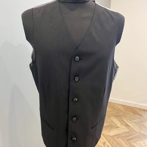 May include: A black vest with a subtle pinstripe pattern, displayed on a mannequin. The vest has a V-neck, a button-up front, and two front pockets. The vest is a formal garment, suitable for layering with a suit or worn on its own.