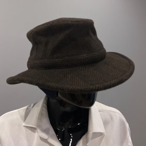 May include: A dark brown bucket hat featuring a subtle herringbone pattern throughout the fabric. The hat has a structured crown and a downward-sloping brim that encircles the head. The material appears textured with a woven finish.