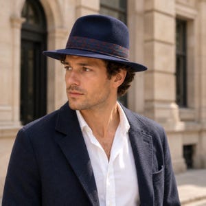 Könnte beinhalten: Ein dunkelblauer Fedora-Hut mit breiter Krempe und einem dekorativen Band, das ein dezentes rot-blaues Muster aufweist. Der Hut wird mit einem weißen Hemd mit Kragen und einem dunkelblauen Blazer kombiniert, was für ein strukturiertes Erscheinungsbild sorgt.