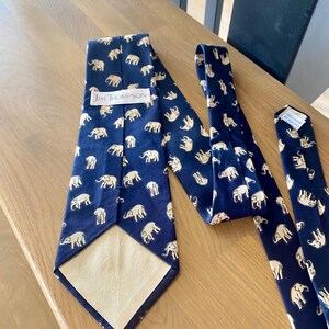 Op de afbeelding: Marineblauwe stropdassen met een herhaald patroon van witte olifanten. E&eacute;n stropdas is volledig weergegeven, terwijl twee andere gedeeltelijk zichtbaar zijn. Het label "Jim Thompson" is zichtbaar op &eacute;&eacute;n stropdas.