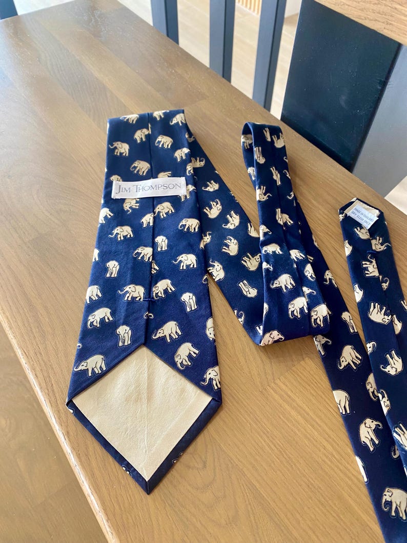 Op de afbeelding: Marineblauwe stropdassen met een herhaald patroon van witte olifanten. E&eacute;n stropdas is volledig weergegeven, terwijl twee andere gedeeltelijk zichtbaar zijn. Het label "Jim Thompson" is zichtbaar op &eacute;&eacute;n stropdas.