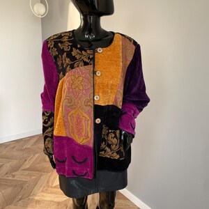 May include: A multi-coloured patchwork jacket with a round neckline and gold buttons. The jacket features panels of purple, gold, black, and pink fabrics. The sleeves are long, and the jacket is made of a velvet-like material. A black leather skirt is also visible.