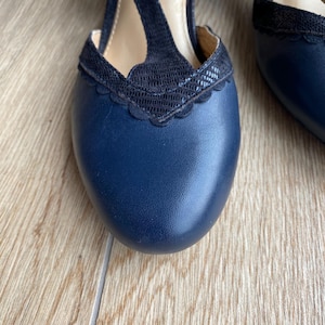 May include: A pair of navy blue T-strap heels featuring a smooth leather toe box and a black textured overlay with a scalloped edge. The design includes a central vertical strap connecting to the vamp, a rounded toe, and a low block heel.