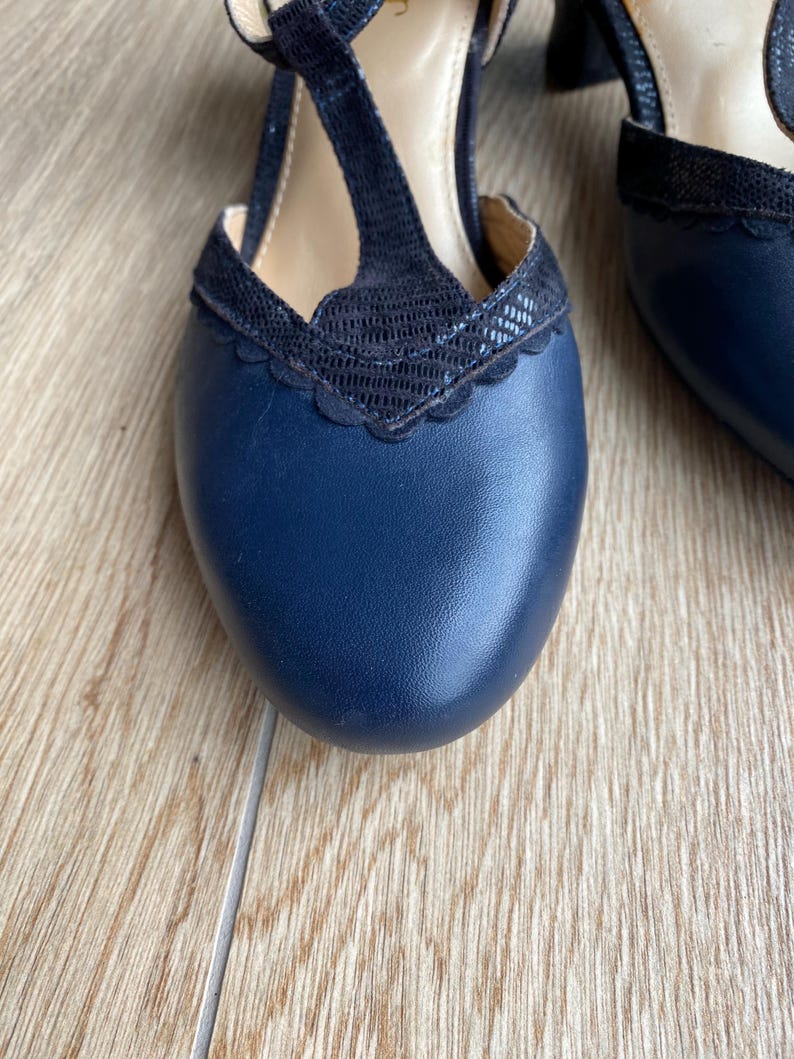 May include: A pair of navy blue T-strap heels featuring a smooth leather toe box and a black textured overlay with a scalloped edge. The design includes a central vertical strap connecting to the vamp, a rounded toe, and a low block heel.