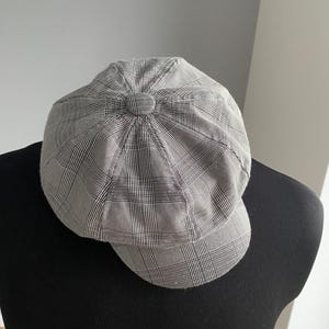 May include: A gray and white plaid newsboy cap with a rounded crown and a small, curved brim. The cap is made of a woven fabric and features a button detail at the top. The cap is displayed on a black mannequin.
