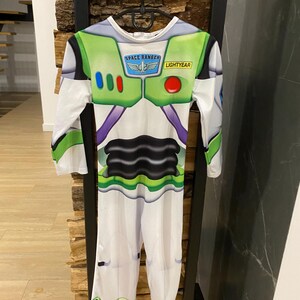 May include: A Buzz Lightyear costume with long sleeves and pants. The costume is primarily white with green, purple, and black accents. The chest area features the text "SPACE RANGER" and "LIGHTYEAR". The costume is hanging on a black hanger.